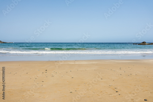 Foto Small dark pebbles dot golden sand as calm turquoise waves roll ashore beneath a clear blue sky, Los Molinos, Fuerteventura, Canary Islands, Spain