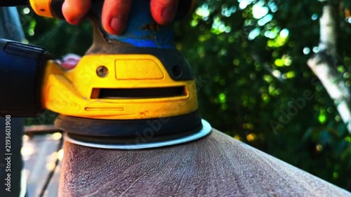Close-up shot of a person using a yellow power sander to smooth a wooden railing or beam. 