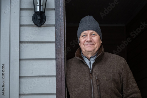 Senior man in warm clothes standing in the doorway of a country house. Useful for mature lifestyle, home ownership, retirement and winter living concepts.