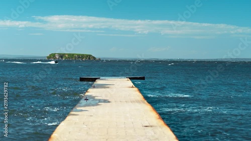 A seaside scene featuring a pier, blue water, and a jet ski cutting through the waves, sending up spray.