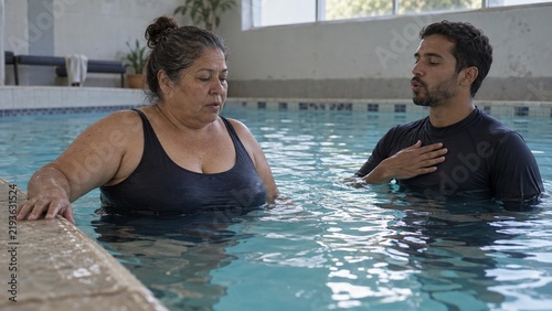 A woman in a black swimsuit stands in a pool, while a man in a black shirt stands opposite her with his hand on his chest, both in an indoor pool area