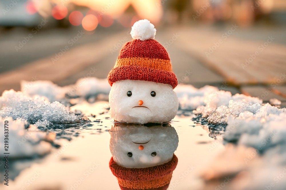 Obraz premium Sad melting snowman head in a red knit hat sitting in a water puddle with reflection on a city sidewalk during winter thaw or early spring season