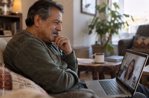 Mature man sitting on sofa having video call with female therapist on laptop. Cozy living room with plant and coffee mug in warm light.
