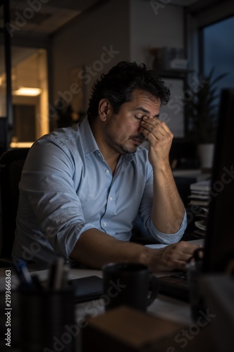 Tired man rubbing his eyes while working late at office desk. Overworked employee suffers from stress and fatigue in dark evening workplace.