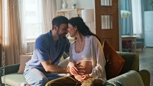 Married couple bonding pregnant belly home sofa. Man touching wife baby bump.