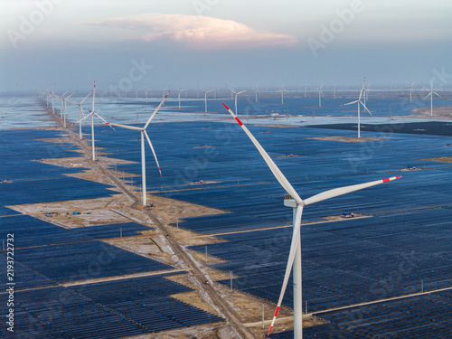 Aerial view of a vast wind and solar farm with rows of wind turbines and solar panels stretching across the landscape under a clear sky in Gujarat, India.