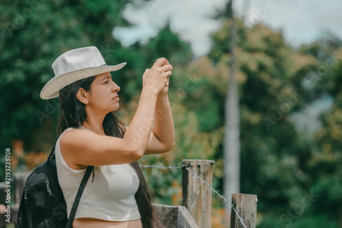 Принти на полотні Woman wearing a white hat and backpack taking a photograph outdoors, enjoying a travel experience surrounded by natural scenery