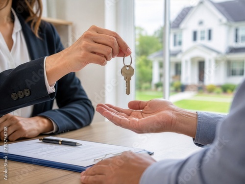 A professional real estate agent hands a house key to a new buyer over a signed contract at a desk.