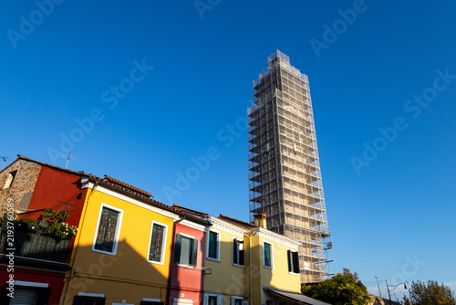 The scaffolded, leaning bell tower undergoes restoration above colorful houses in Burano, Venice, Italy