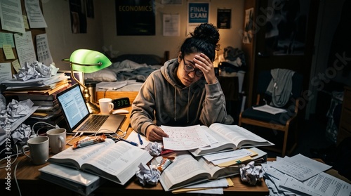 Exhausted female college student holding her head in despair while studying late at night at a messy desk in a dorm room