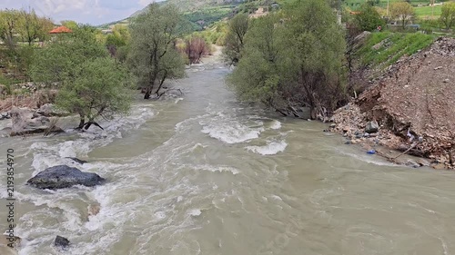 Swift, muddy mountain river winds past lush green trees and hillside houses beneath a partly cloudy sky, framed by rolling green mountains.
📍Dorud village, Sarvabad, Kurdistan 