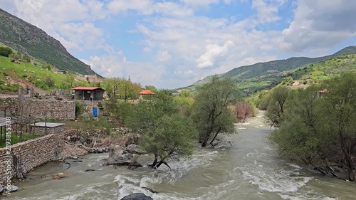 Swift, muddy mountain river winds past lush green trees and hillside houses beneath a partly cloudy sky, framed by rolling green mountains.
📍Dorud village, Sarvabad, Kurdistan 