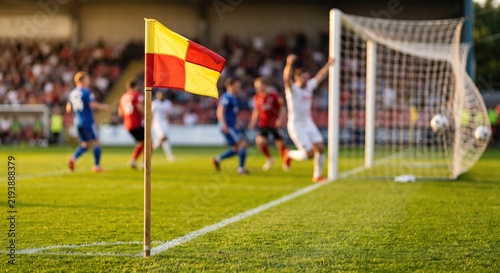 Red and yellow corner flag on a green soccer pitch with a blurred background of players celebrating a goal in the net