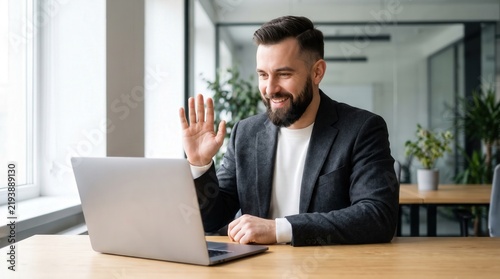 Professional bearded businessman waving at laptop during virtual meeting, capturing friendly remote communication for corporate marketing and digital collaboration software.