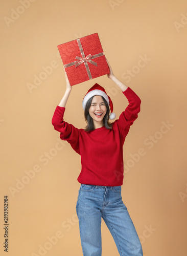 Cheerful Asian woman celebrating Christmas wearing a santa hat and carrying a red gift box on the head while smiling.