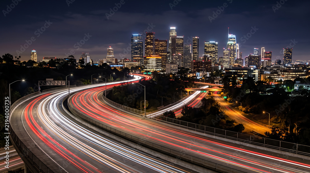 Fototapeta premium Curved Highway Light Trails Leading Toward Illuminated Downtown City Skyline at Night Long Exposure View