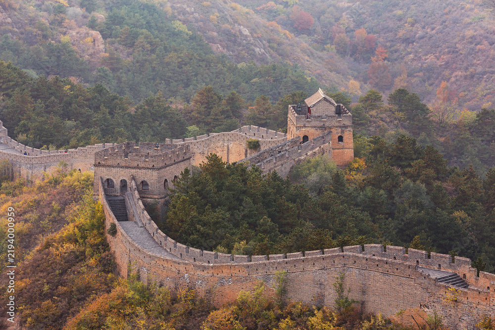 Fototapeta premium Ancient stone fortification winds across autumn mountain slopes, flanked by dense forests with seasonal foliage.