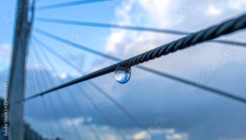 A single clear water droplet hanging from a steel cable on a bridge.