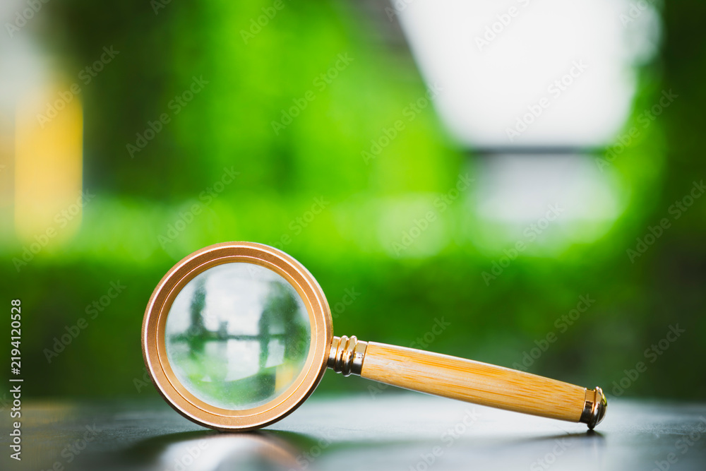 Obraz premium Search and discovery concept, vintage magnifying glass with wooden handle on reflective surface with blurred green nature background, representing audit, research, inspection, and analysis.