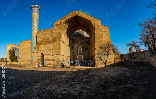 Side view of the Amir Temur Mausoleum, also known as Gur-e Amir, in Samarkand, Uzbekistan. The scene shows the weathered brick entrance portal, a tall turquoise-tiled minaret, the ribbed dome rising b