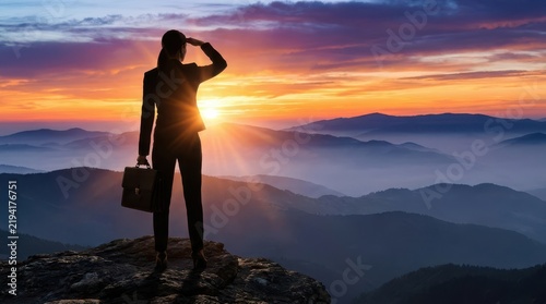 Ambitious businesswoman standing on a mountain peak holding a briefcase looking forward at sunrise representing success leadership and future vision