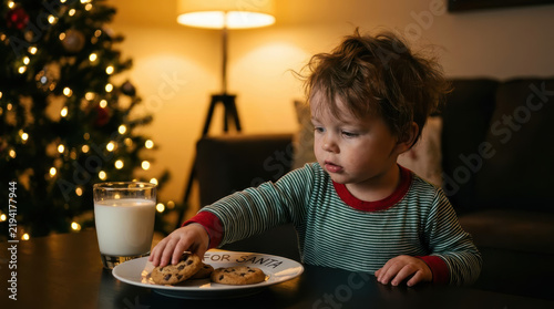 Toddler playfully stealing chocolate chip cookie beside milk glass at warmly lit holiday table, expressing curiosity and delight during cozy seasonal family moment