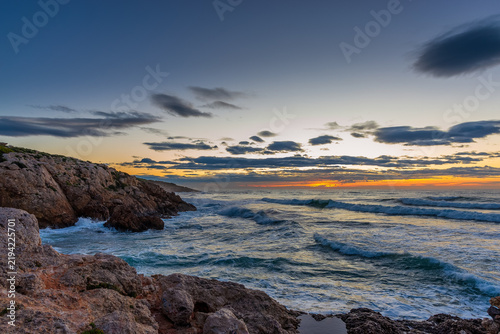 Dramatic sunrise over a rocky Mediterranean coastline with turquoise waves crashing against limestone cliffs under a blue and orange sky with scattered clouds, peaceful seascape at dusk in Europe