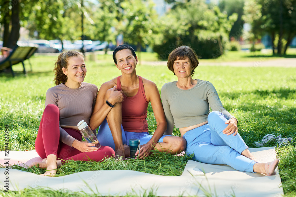 Fototapeta premium Portrait of young adult and middle aged Caucasian women sitting on exercise mats in park resting after fitness workout. Imagery suitable for wellness apps, healthy lifestyle campaigns