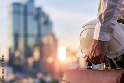 A business professional holding a white hard hat and leather briefcase with a blurred city skyline at sunset, urban development and corporate real estate investment concept