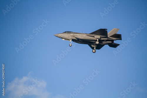 F-35A Lightning II fighter aircraft flying across a blue sky with wheels down, photographed in side profile showing its distinctive stealth aircraft design.