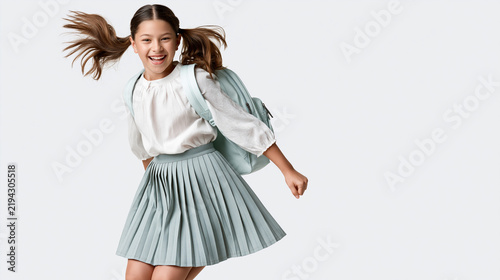 Asian girl with pigtails wearing a white blouse and pleated skirt jumps joyfully while carrying a light blue backpack against a plain background