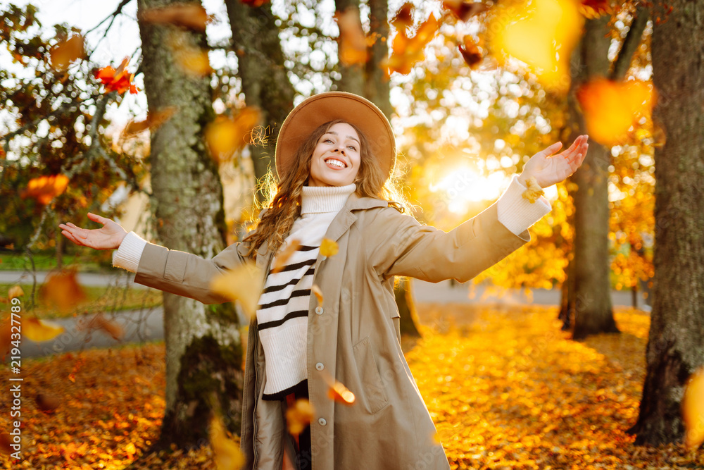Obraz premium Woman wearing hat and trench coat is having fun in park covered with colorful leaves. Young woman enjoys autumn woods and tosses yellow leaves into air. Concept of leisure, weekends, and autumn.