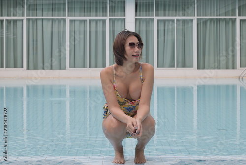 A smiling Asian woman in a yellow floral swimsuit crouches at the edge of a swimming pool