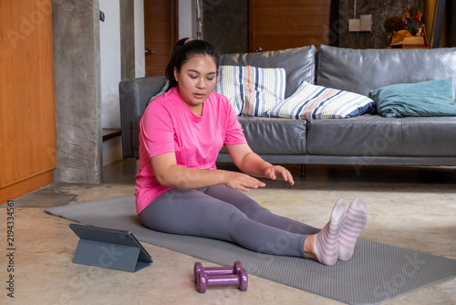 Woman performing fat burning exercise on yoga mat at home with tablet and dumbbell nearby