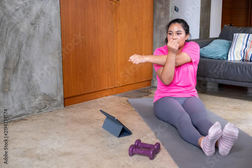 Fat woman doing exercise on mat with dumbbell and tablet at home while stretching her legs and looking focused during workout session