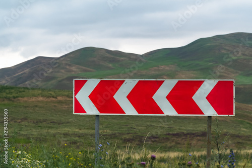  Red and white chevron road sign against green mountains in Kyrgyzstan.