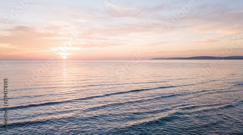 Beautiful pink and golden sunset over calm ocean waves with soft clouds and distant coastline mountains during peaceful evening reflection