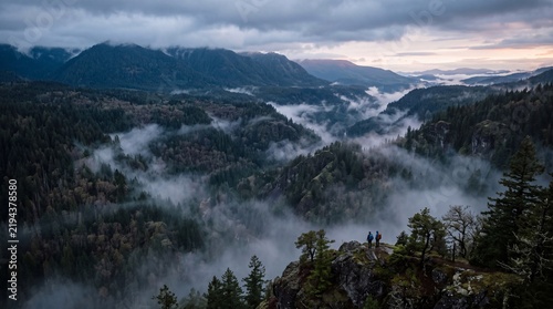 Two adventurous hikers standing on rocky cliff edge overlooking majestic misty mountain valley forest during moody dawn sunrise scenery