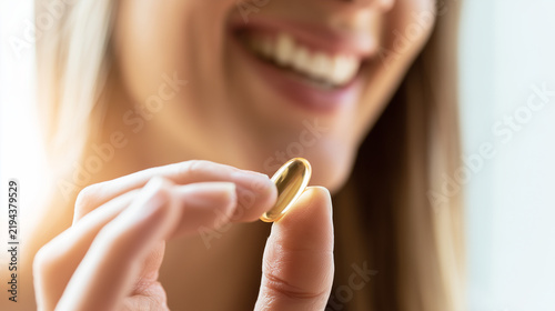 Young woman holding a translucent golden softgel capsule close to her mouth, smiling before taking a daily dietary supplement in bright natural light, vitamin capsule, nutritional