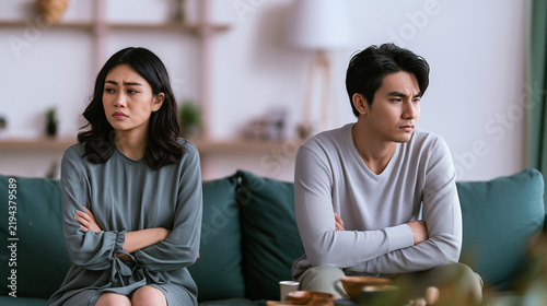 Young Asian husband and wife sitting apart after an argument, woman showing anger while the man reacts with frustration and avoids eye contact in their home interior, marriage prob