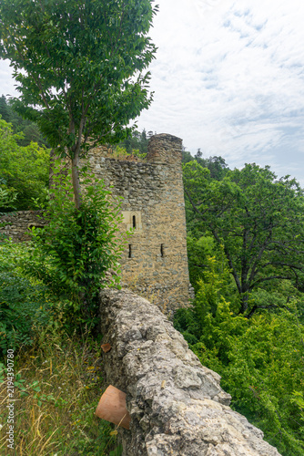 A medieval tower with narrow windows against a high wall. A forest and sky with clouds in the background. Amlivi, Amlevi complex. Kvemo Kartli, Tetritskaro, Georgia