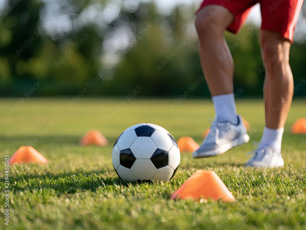 Obraz premium Soccer player training with cones and ball on grass field. Close-up of legs, football, and training equipment outdoors on a sunny day.