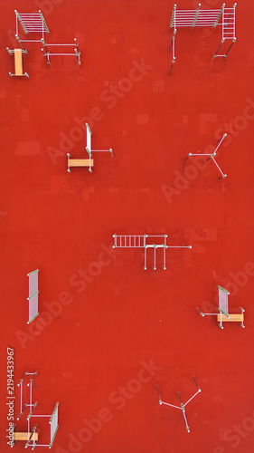Aerial view of a modern outdoor calisthenics park with metal exercise equipment and benches set against a vibrant red rubberized ground surface in Riga, Latvia.