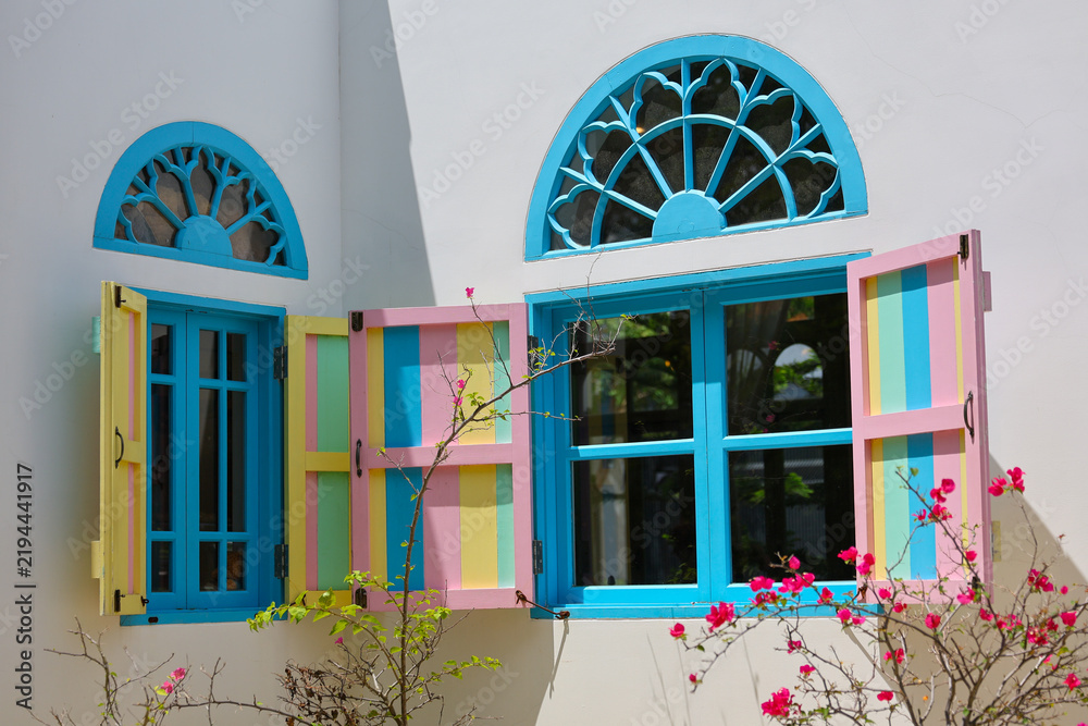 Obraz premium Colorful seaside house facade with turquoise windows, arched glass fanlights, open pastel shutters, and flowering bougainvillea creating cheerful exterior