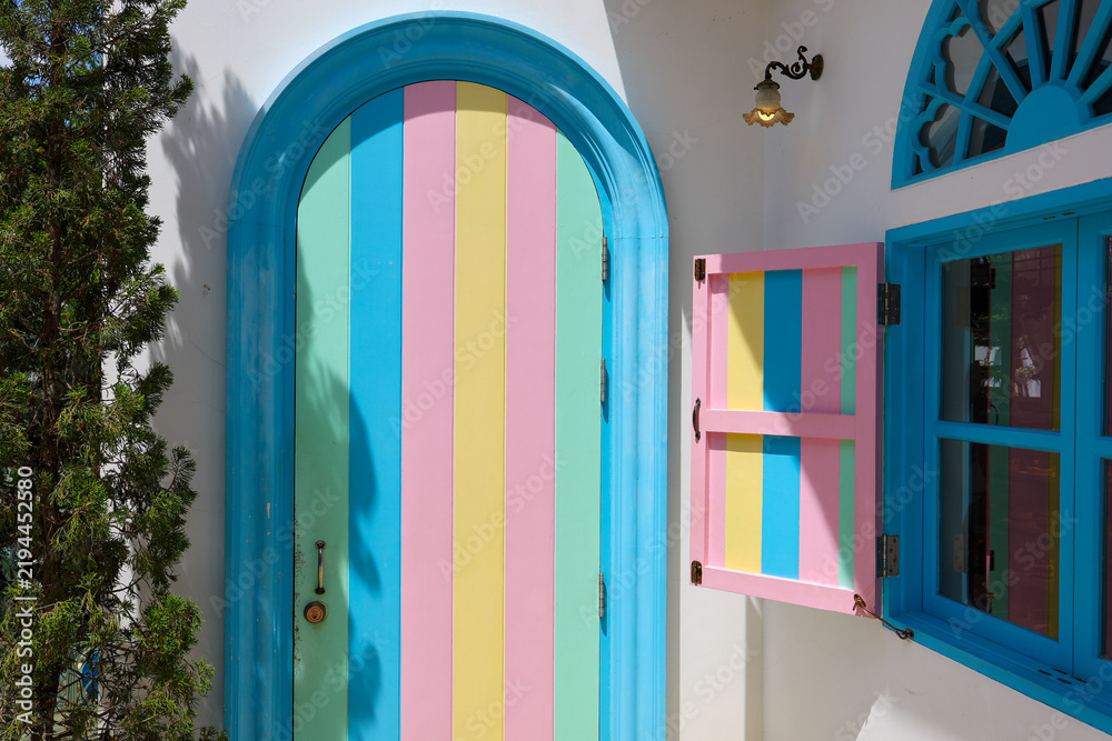 Obraz premium Turquoise arched door and colorful shutter on white Mediterranean house exterior create cheerful geometric facade with vivid pastel stripes and strong sunlight