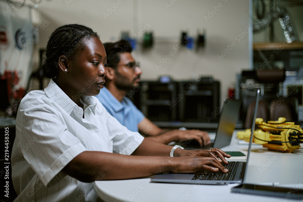 Fototapeta premium African American young woman typing on laptop in engineering workshop alongside colleague. Useful for IT software development branding and STEM education