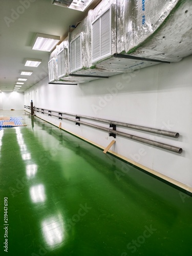 A perspective view of a spacious industrial cleanroom hall featuring a glossy green epoxy floor, polished stainless steel process piping mounted along clean white sandwich panel walls, overhead air ha
