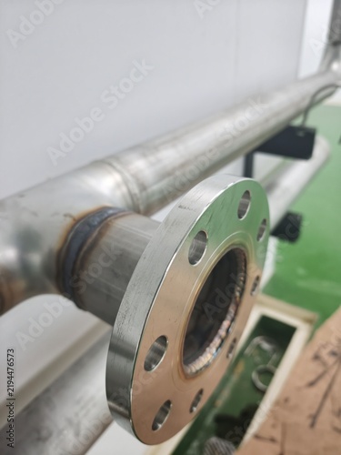 A close-up shot of a precision-welded stainless steel pipe flange with bolt holes, installed on an industrial process pipeline against cleanroom walls and epoxy flooring.