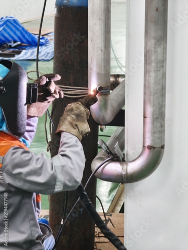 A close-up shot of a professional welder wearing a flip-up protective welding mask and heavy leather gloves, holding a TIG torch and filler rod to perform precise welding on a stainless steel pipe 