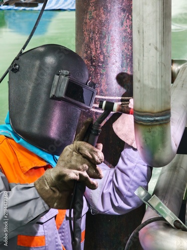 A close-up shot of a professional welder wearing a flip-up protective welding mask and heavy leather gloves, holding a TIG torch and filler rod to perform precise welding on a stainless steel pipe 
